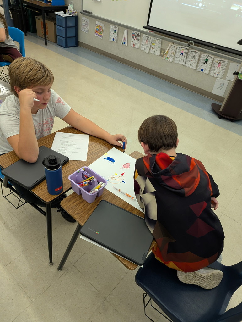 Two students sit at a classroom desk working together on a drawing activity using markers and crayons. One student leans in to color while the other watches thoughtfully, with a basket of art supplies and Chromebooks on the desk, highlighting collaboration and creativity during a club activity.
