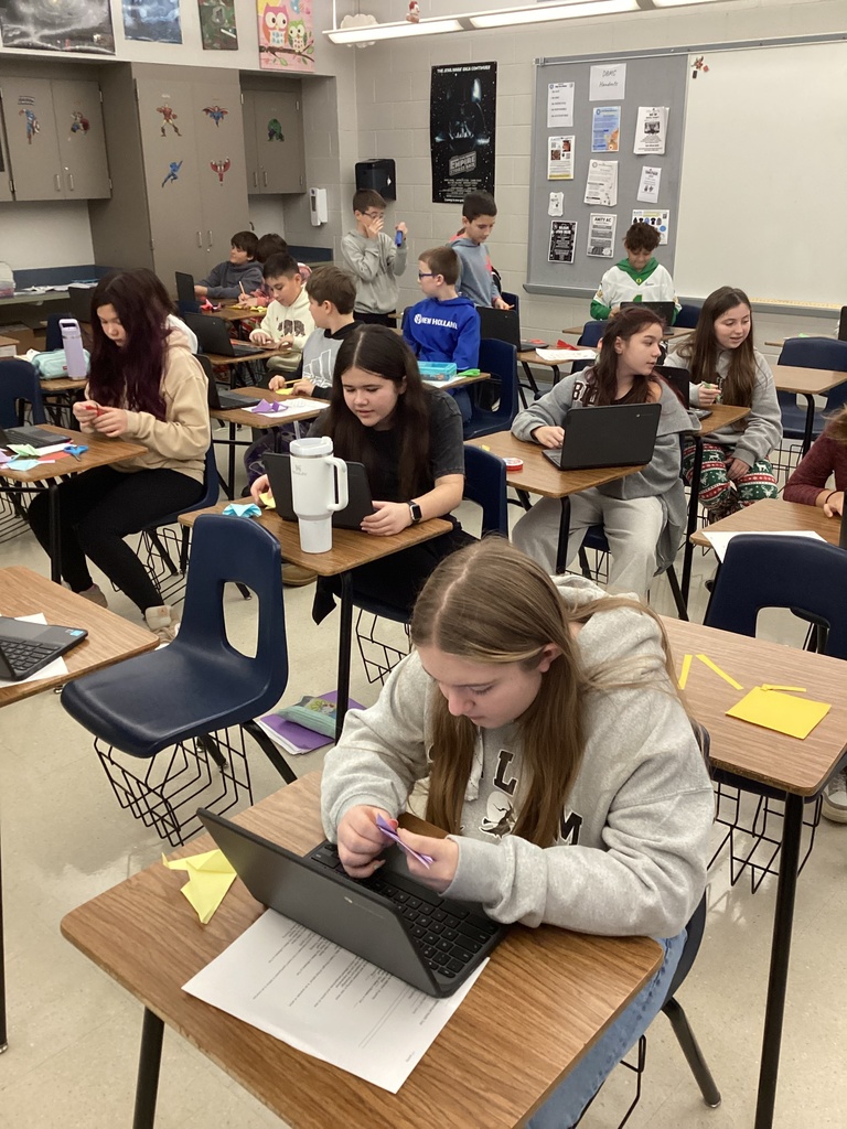 Sixth-grade students work independently and in small groups at classroom desks, folding paper and using Chromebooks during a hands-on club activity. Paper creations, laptops, and classroom materials are visible throughout the room, highlighting student focus, creativity, and engagement.