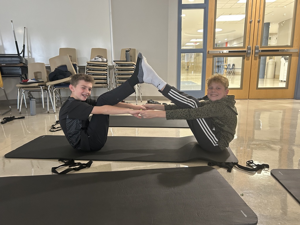 Two students sit facing each other on exercise mats, smiling as they balance and stretch with their feet raised and hands clasped during a yoga or movement club activity. Stacked chairs and a school hallway are visible in the background, highlighting teamwork and focus.