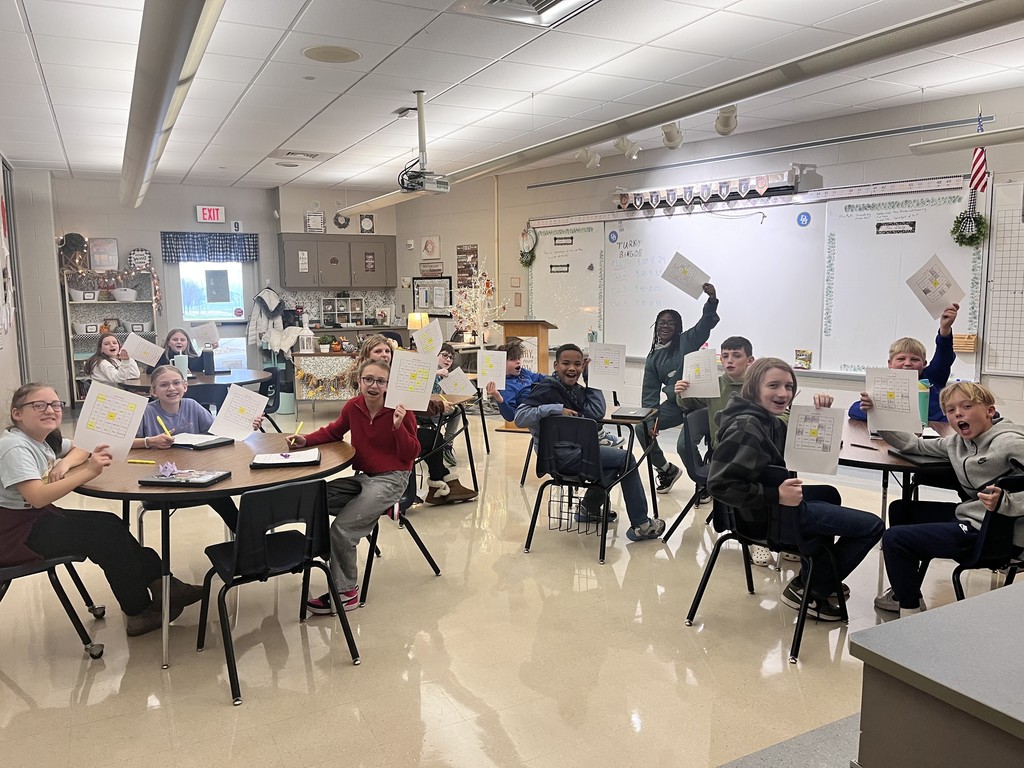 Sixth-grade students sit at round tables in a classroom, smiling and holding up completed activity sheets during a club rotation. The bright classroom includes desks, a whiteboard, and classroom decorations, capturing students’ excitement and engagement as they proudly show their work.
