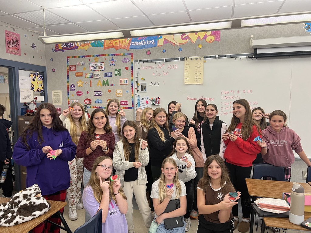 A group of smiling sixth-grade students stand together in a classroom, holding up small handmade craft items they created during a club activity. The classroom walls are decorated with colorful posters and a whiteboard, and desks are arranged around the room as students pose proudly with their projects.