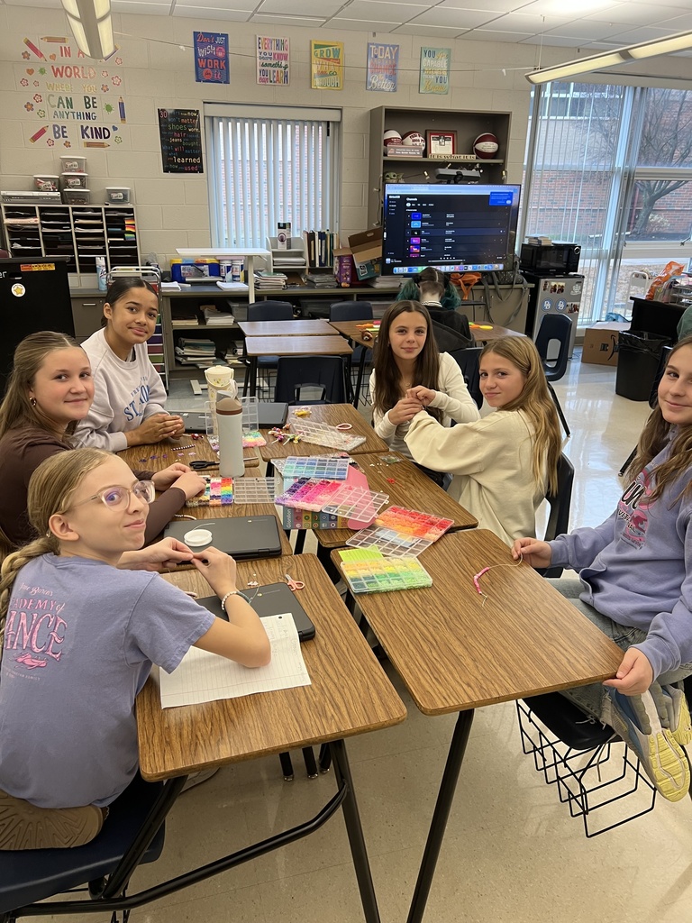 A group of sixth-grade students sit together at classroom tables, smiling and working on bead craft projects during a club activity. Colorful bead organizers, string, and partially completed bracelets are spread across the tables, showing students collaborating and enjoying hands-on creativity in a welcoming classroom setting.