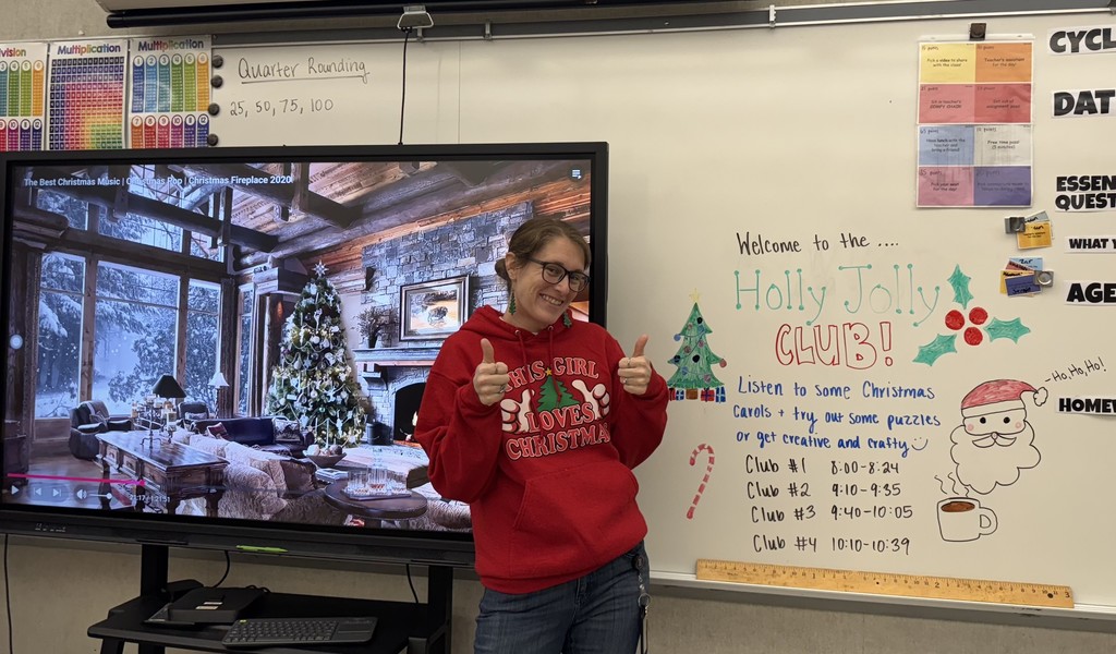 A teacher stands smiling in front of a classroom whiteboard decorated for the “Holly Jolly Club,” giving two thumbs up. Beside her, a large screen displays a cozy winter scene with a Christmas tree and fireplace, while the whiteboard lists club activities and schedule with festive drawings like holly, candy canes, and a Santa face.
