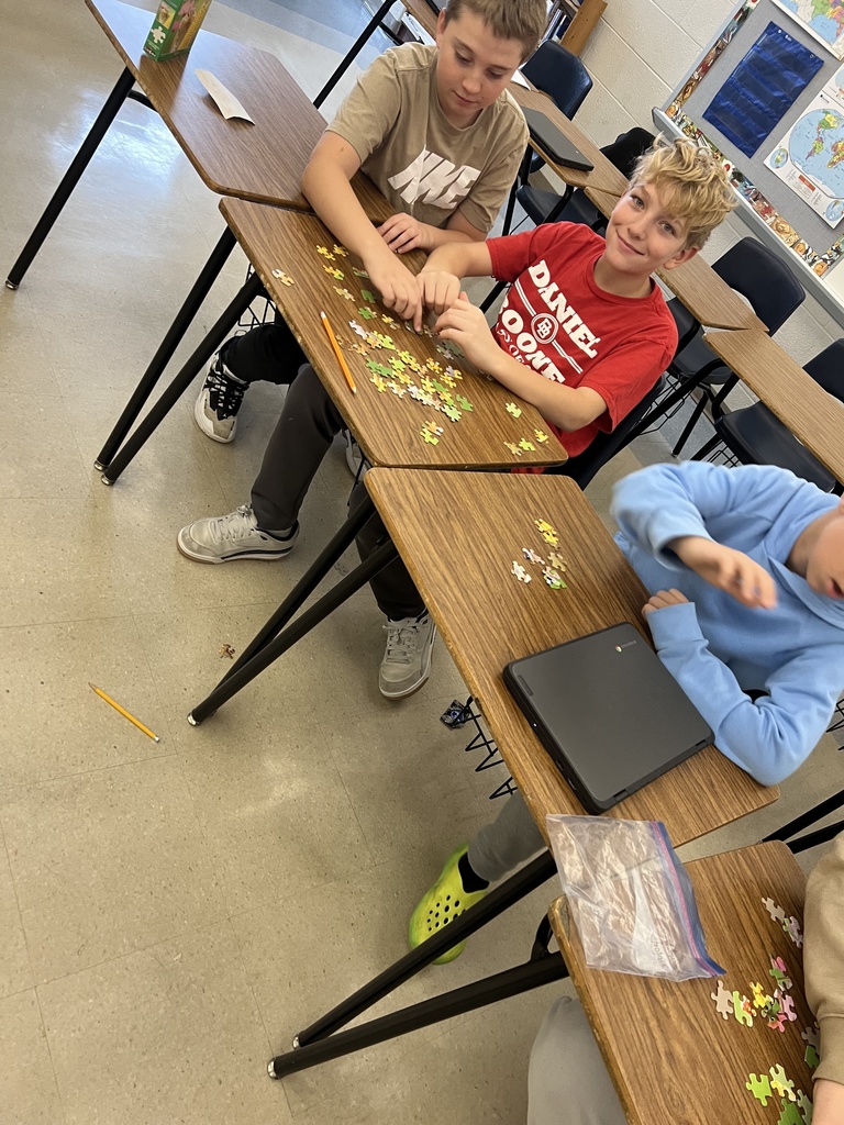 Two sixth-grade students sit at classroom desks, working together to assemble a jigsaw puzzle spread across the tabletop. Puzzle pieces, a pencil, and Chromebooks are visible, capturing a hands-on club activity focused on collaboration, problem-solving, and fun.