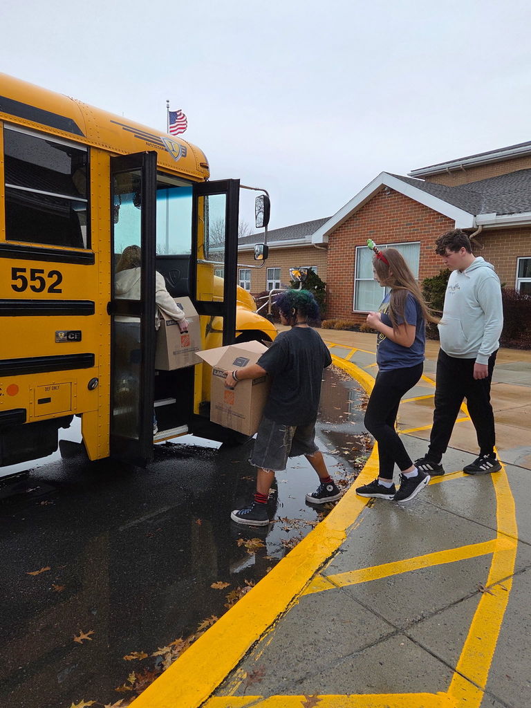 Four high school students load large boxes of donations onto a yellow school bus on a rainy day. One student lifts a box onto the bus while the others wait in line, standing near a brick school building with an American flag flying above.