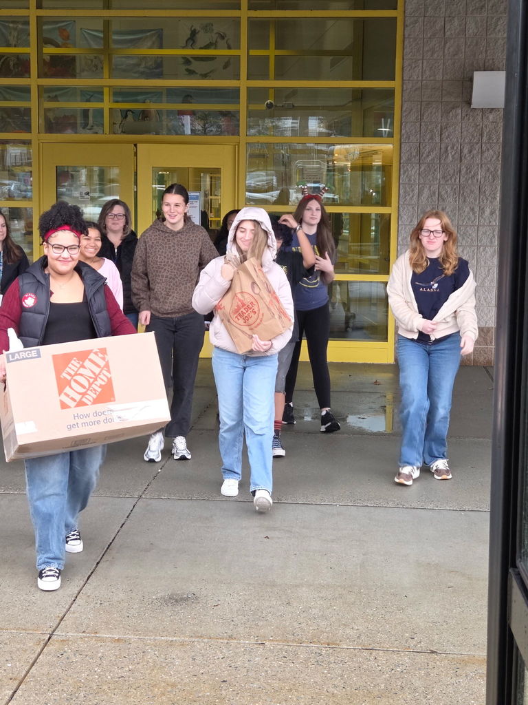 A group of high school students exit a school building with bright yellow-framed doors, smiling as they carry donations. One student in front holds a large Home Depot box, while another carries a Trader Joe’s paper bag. Others follow behind, some wearing festive accessories.