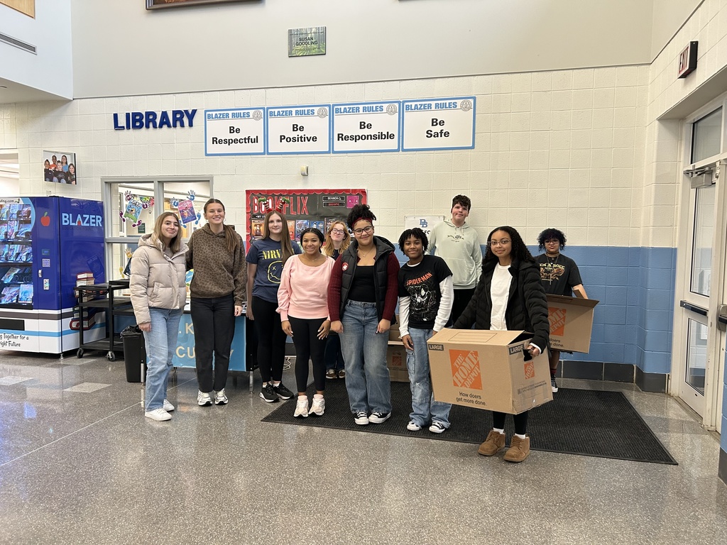 A group of high school students stand inside a school lobby near the library, posing with smiles and large donation boxes. Behind them are signs displaying the “Blazer Rules” — Be Respectful, Be Positive, Be Responsible, Be Safe — and a vending machine labeled “Blazer.”