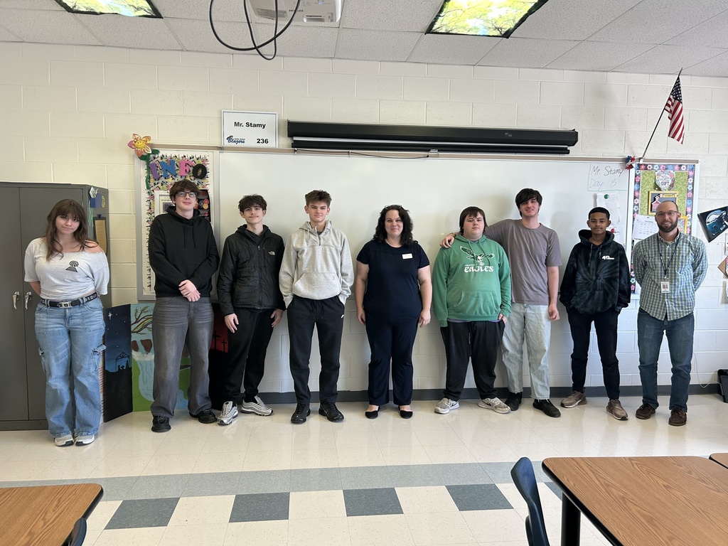A group of high school students and two adults stand in a classroom lined up in front of a whiteboard. Kristen Gates from Tompkins Community Bank stands in the center, flanked by students from the DBAHS Entrepreneurship class and their teacher. Everyone is smiling after the guest speaker session.