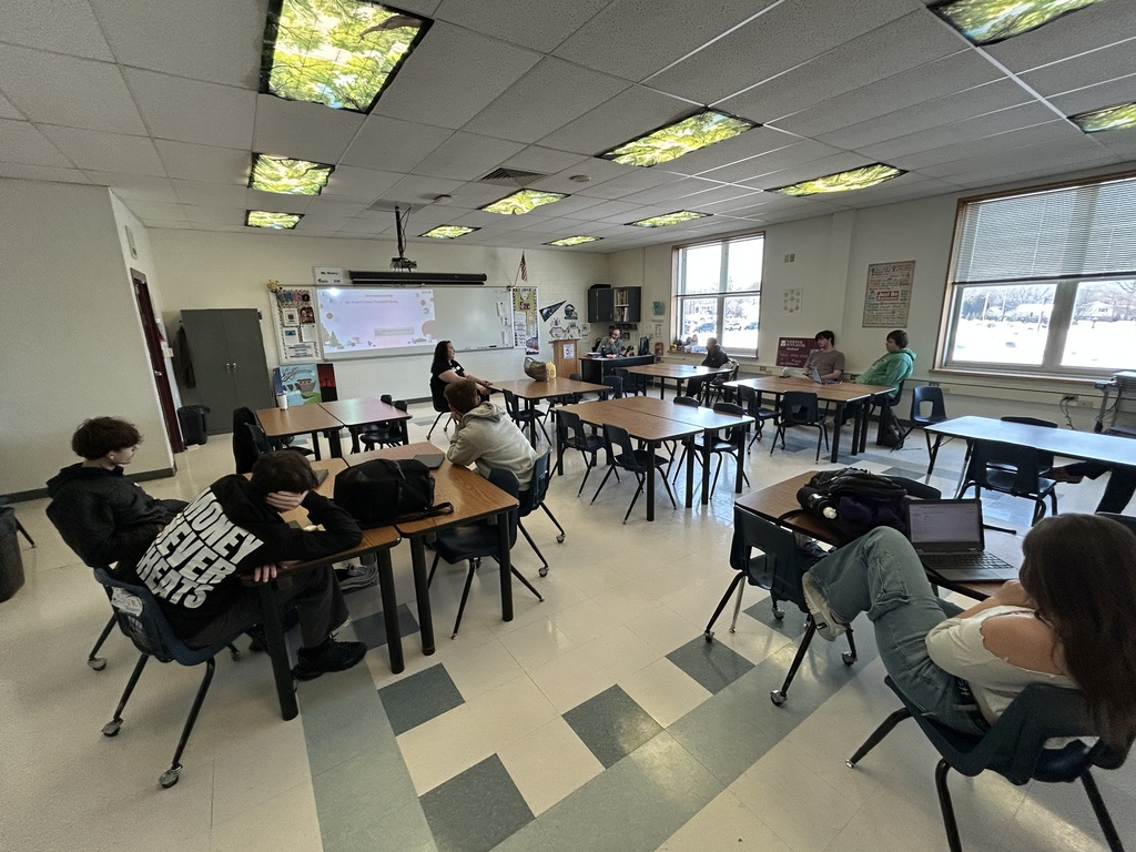 Students sit at desks in a high school classroom listening to a guest speaker at the front of the room. A presentation is projected on the whiteboard behind the speaker, and natural light streams in through large windows. The classroom setup reflects a relaxed, discussion-based environment.