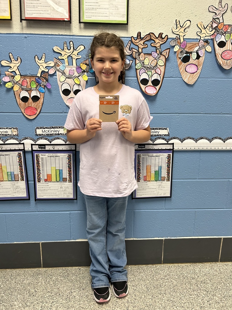 A smiling elementary student stands in front of a decorated classroom wall holding a $100 Amazon gift card.