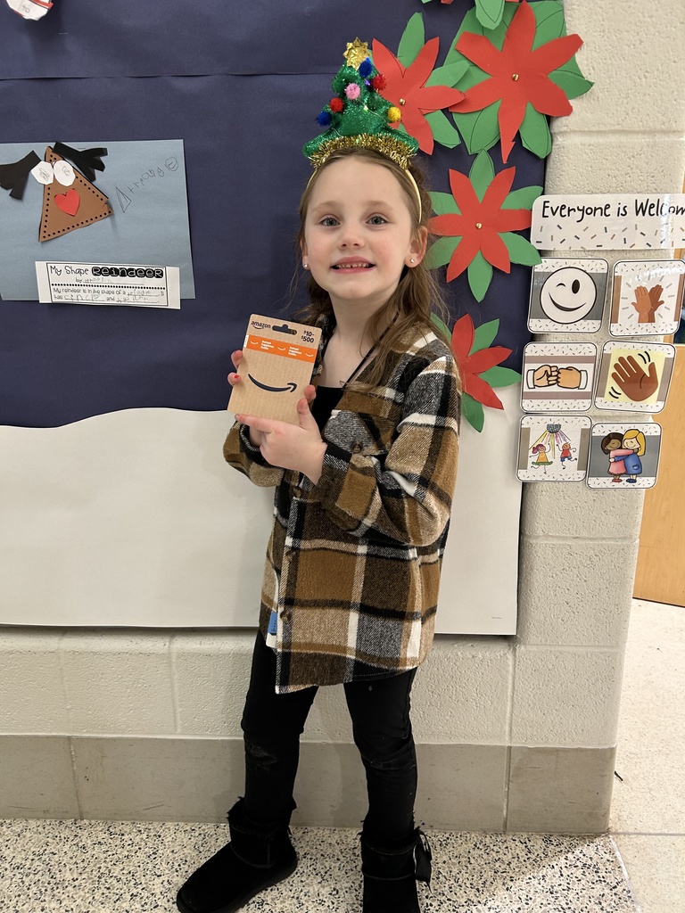 A smiling elementary student stands in front of a decorated classroom wall holding a $100 Amazon gift card.