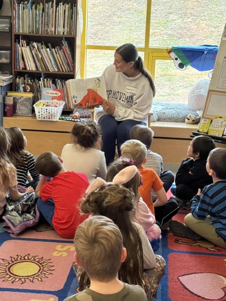 High School Student reading to a class of students at the primary center. Student is sitting on a rocking chair while students are seated on a colorful alphabet carpet.