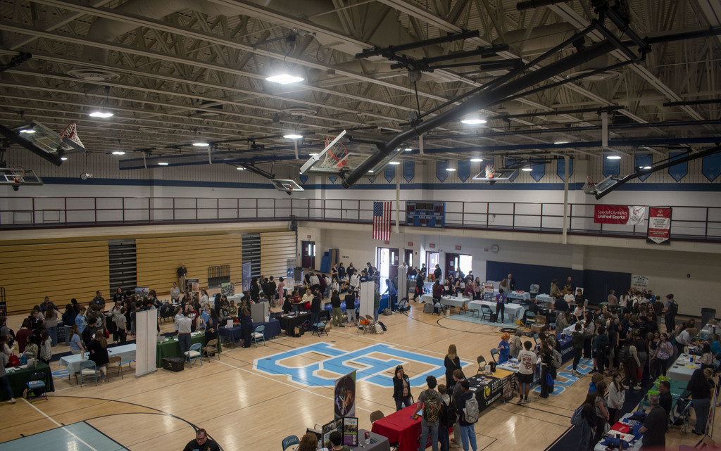 Picture of our gym with the DB Logo in the the floorboards. Tables are setup in a u shape around the perimeter with businesses representing their companies talking to kids.