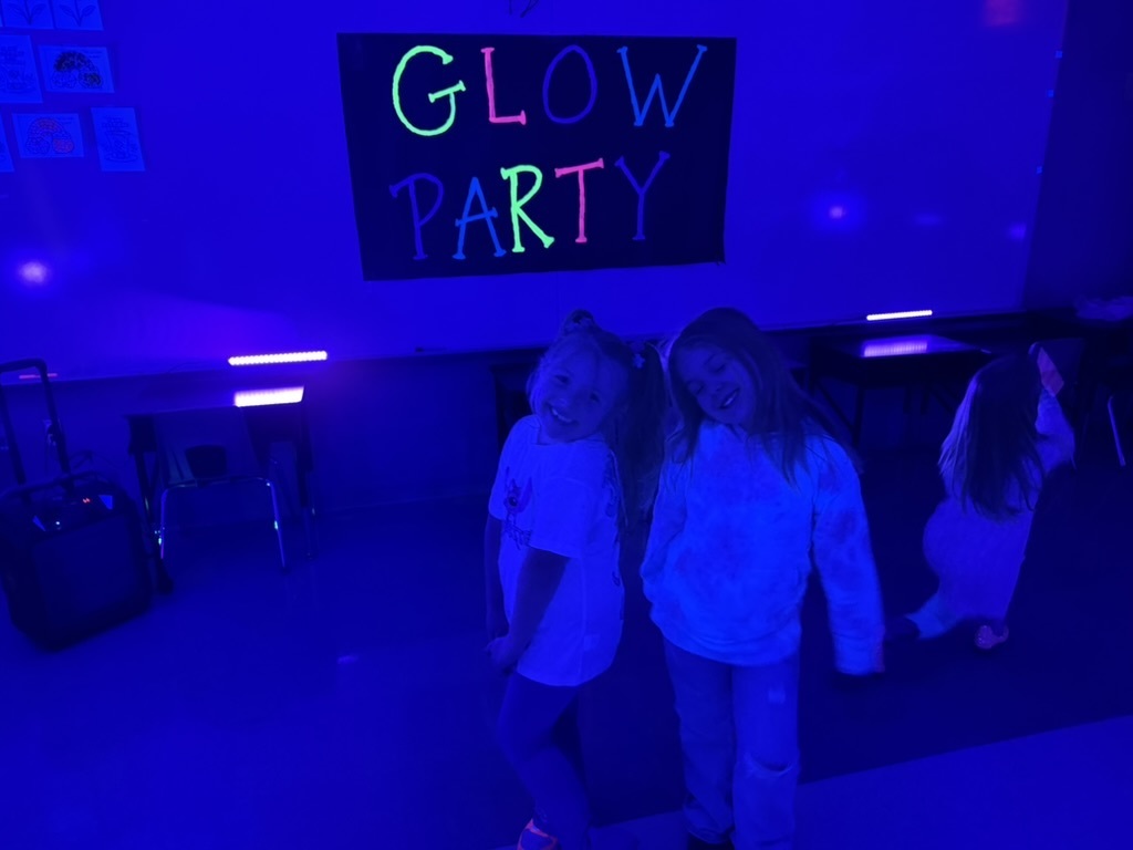 Blacklight picture of two students standing in front of a GLOW PARTY sign.