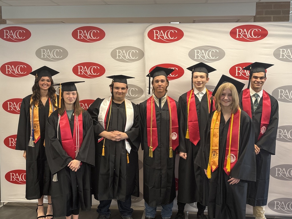 Seven students stand shoulder-to-shoulder in front of a step-and-repeat backdrop with repeating RACC logos. They are wearing black graduation gowns, caps, and red stoles, with some also wearing gold honor cords. The group is smiling and facing the camera in an indoor setting.