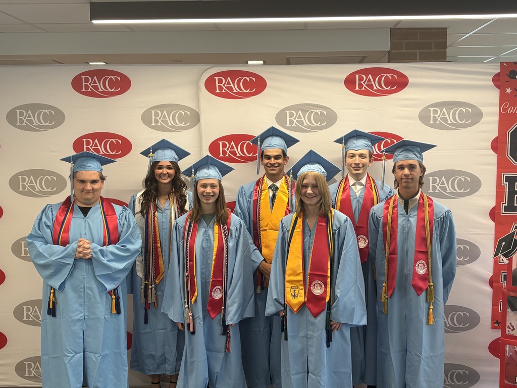 The same seven students pose in front of the RACC logo backdrop, now dressed in light blue graduation gowns and caps. Several are wearing red stoles and gold honor cords. They are standing close together, smiling at the camera in the same indoor location.