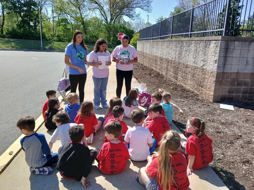 Three high school students standing outside on the sidewalk with 17 kindergarten students sitting on the sidewalk
