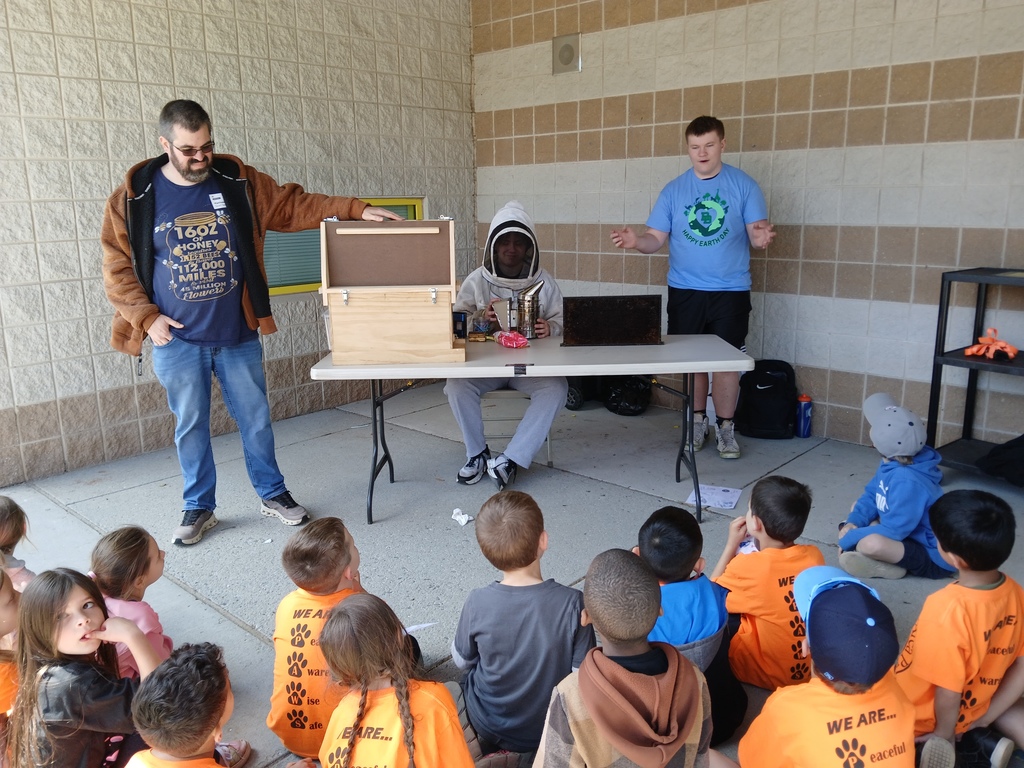 Students sitting on sidewalk listening to a demonstration from a beekeeper. One high school student is speaking to the kindergarteners while one person is sitting at a table wearing a beekeeper outfit. There is another person standing next to a bee box.