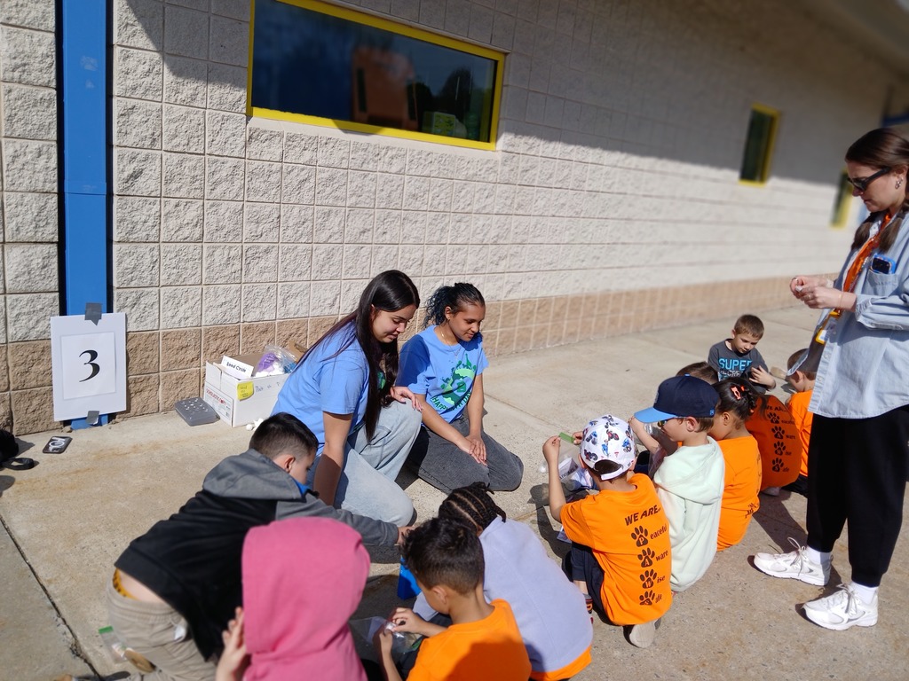 Kindergarten students sitting on a sidewalk learning about planting from two high school students.
