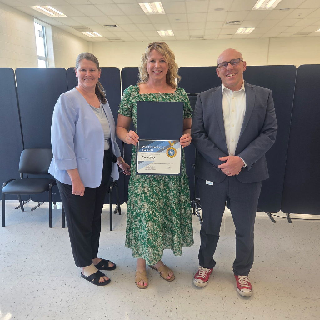 Principal Melanie Hefter standing with School Counselor Carrie Berg and Superintendent Tom Voelker. Carrie is holding a Sweet Impact Award Certificate. The trio are standing in the Daniel Boone Board Room.