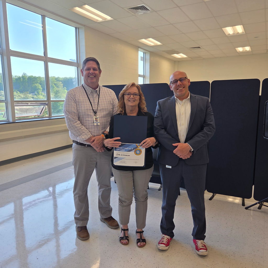 Principal Jim Towers standing with Attendance Secretary Beth Williamson and Superintendent Tom Voelker. Beth is holding a Sweet Impact Award Certificate. The trio are standing in the Daniel Boone Board Room.