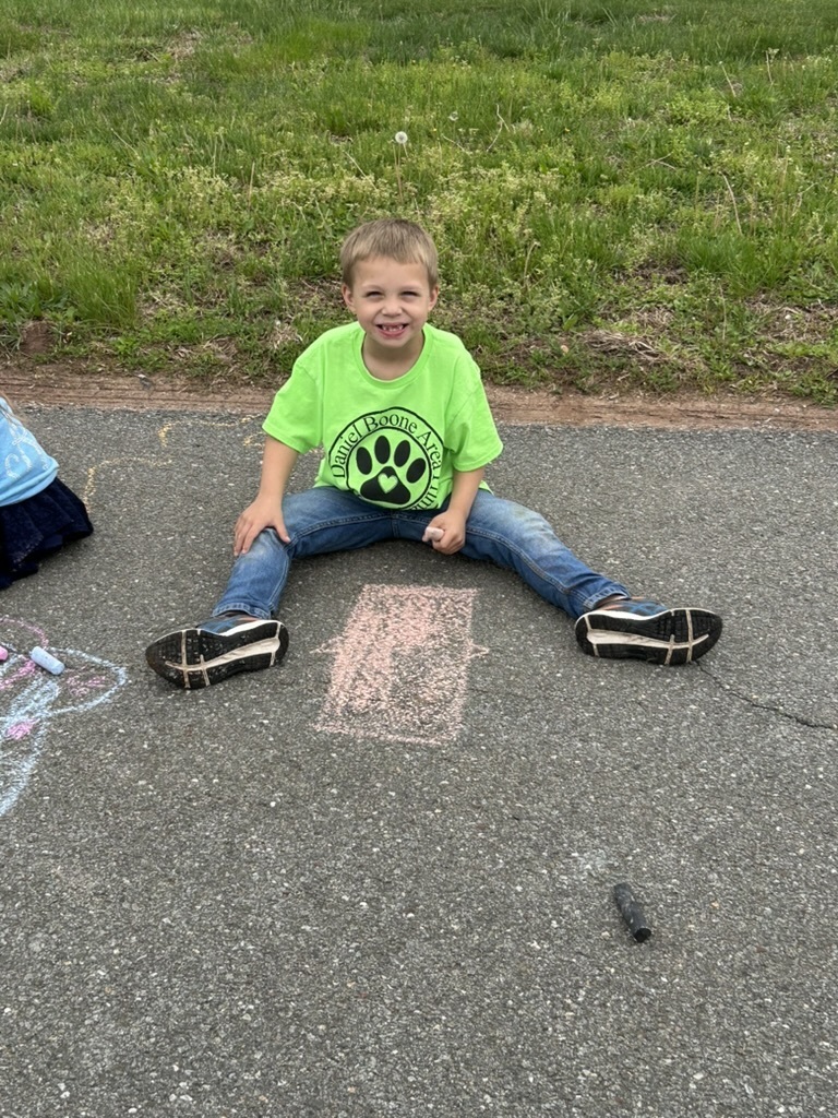 A picture of a student sitting on the playgroundwearing a green shirt holding sidewalk chalk.  He drew a rectangle on the playground macadam.