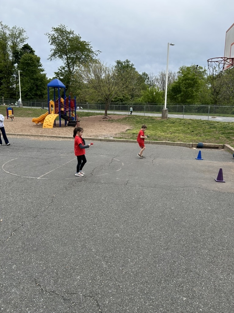 Picture of two students wearing red tshirts on the playground with cones competing in an egg race.