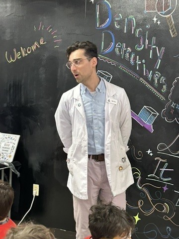 Picture of a man wearing a white lab coat standing in front of a blackboard wall. In chalk is written Welcome Density Detectives with random chalk doodles.