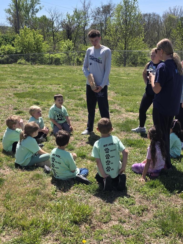 A group of kindergarten students wearing green Daniel Boone Area Primary Center shirts sit in a circle outside on a sunny day, smiling and participating in a teamwork activity led by high school student ambassadors on the grass near the school building.