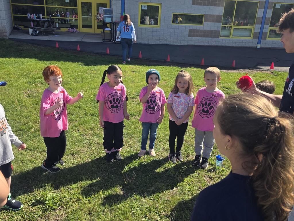 A group of kindergarten students wearing pink Daniel Boone Area Primary Center shirts stand in a circle outside on a sunny day, smiling and participating in a teamwork activity led by high school student ambassadors on the grass near the school building.