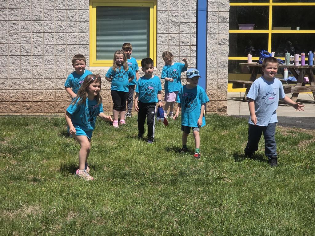 A group of kindergarten students wearing blue Daniel Boone Area Primary Center shirts stand awaiting to start running outside on a sunny day, smiling and participating in a teamwork activity led by high school student ambassadors on the grass near the school building.