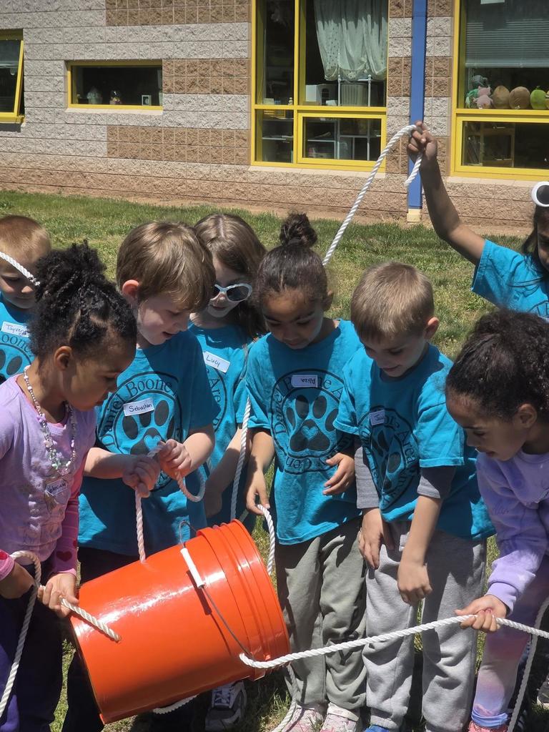 A group of kindergarten students wearing blue Daniel Boone Area Primary Center shirts stand in a circle outside on a sunny day, smiling and participating in a teamwork activity led by high school student ambassadors on the grass near the school building.