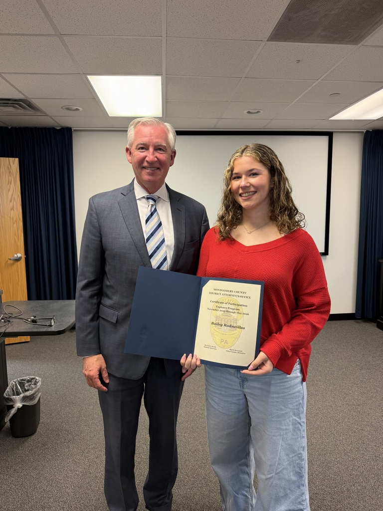 Picture of student wearing a red sweater and jeans holding a certificate of participation in a boardroom standing beside a Montgomery County District Attorney wearing a grey suit and blue striped tie.