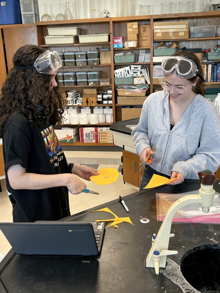 Two girls wearing goggles on their head cutting yellow felt at a lab table.