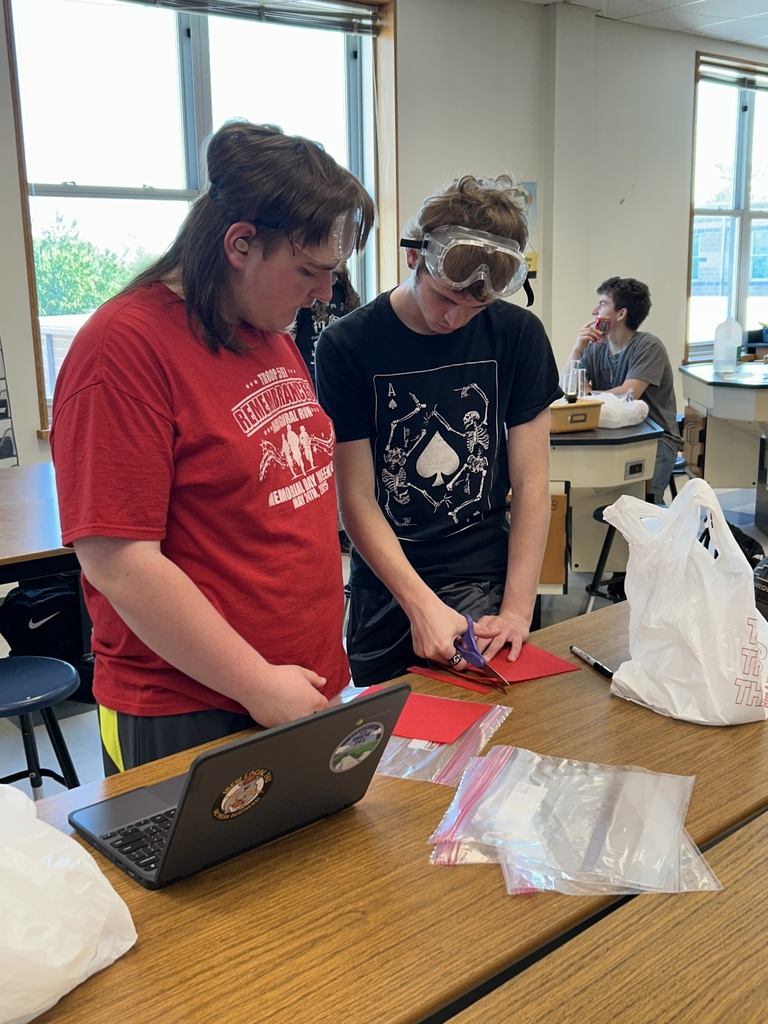 Two boys standing at a lab table wearing goggles on their head cutting red felt.