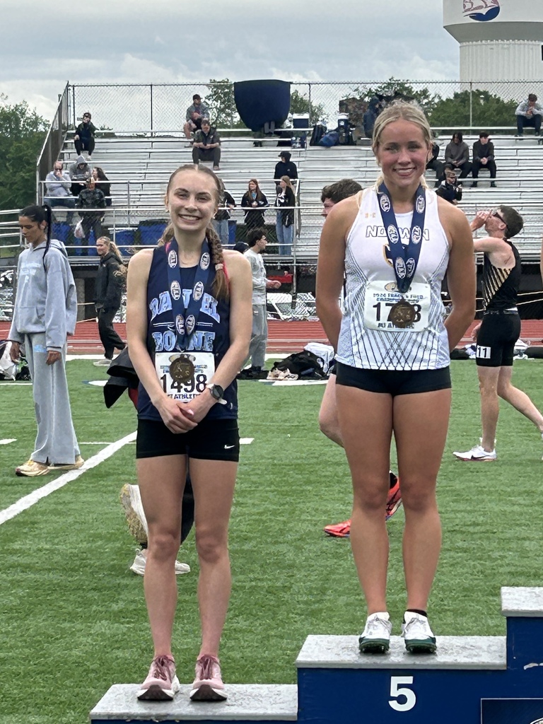 Two girls wearing medals and their track uniforms standing on the podium in the middles of the football field.