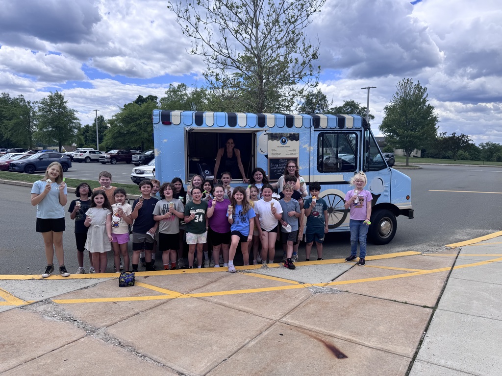 Mrs. Kern's class standing in front of the Sweet Ride Ice Cream truck holding their ice cream cones outside on the curb of the parking log with cloudy blue skies , trees and cars in the background.