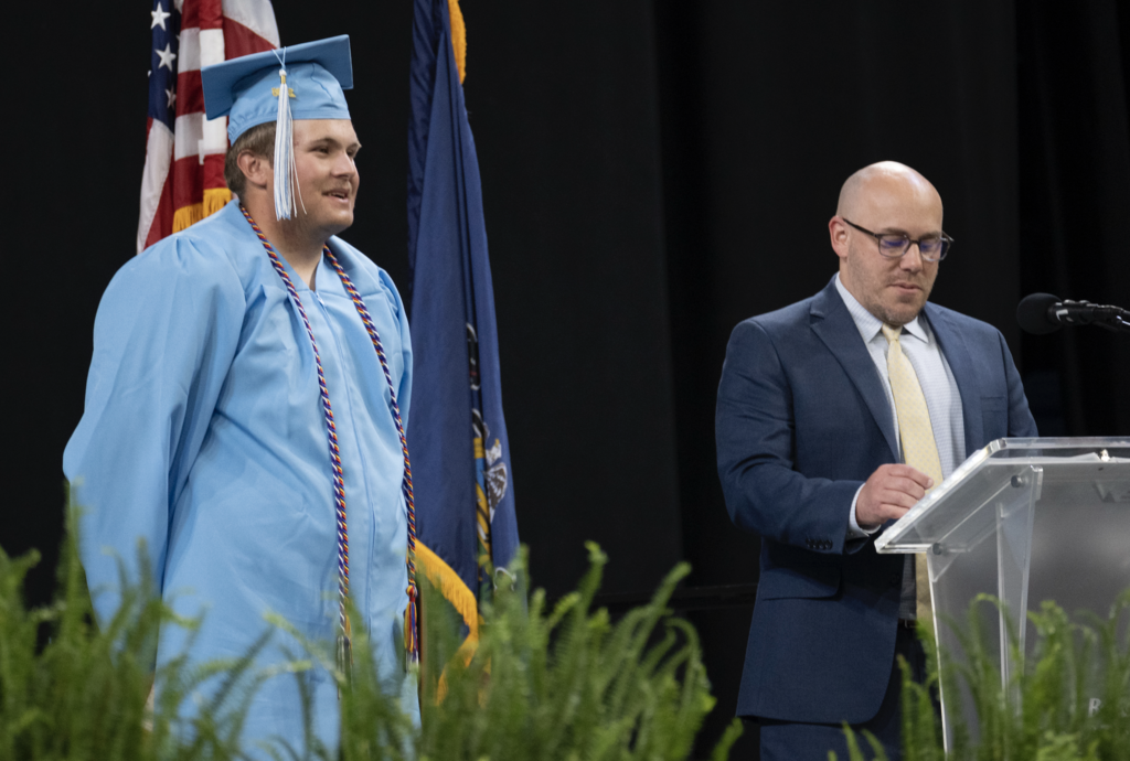 Owen wearing his light blue cap and gown with tassel and cords standing in front of an American and Pennsylvania flag. Next to him is Superintendent Tom Voelker reading from his notes at a podium.