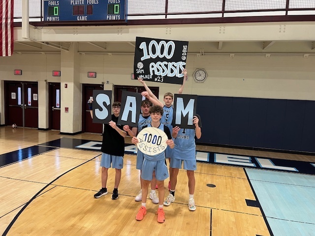 Five Daniel Boone boys volleyball players stand together in the high school gym celebrating teammate Sam Walker’s achievement of reaching 1,000 career assists. Three teammates hold large letters spelling “SAM,” while another holds a sign overhead that reads “1,000 Assists – Sam Walker #23.” Sam stands in front holding a volleyball decorated with “1,000 assists.” The group is dressed in team practice attire and poses on the gym floor in recognition of the milestone.