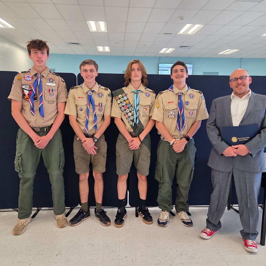 Four students wearing their scout uniforms stand with the superintendent in the board room being recognized for their Eagle Scout awards