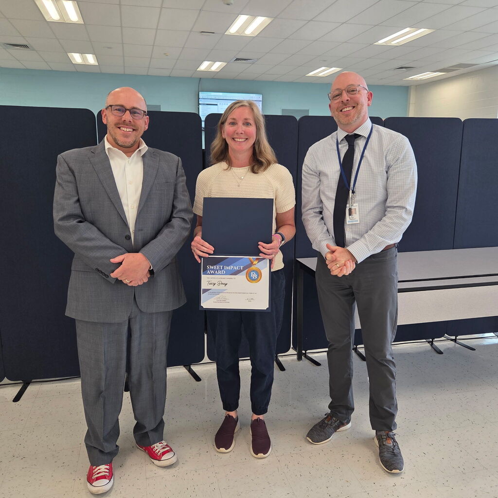 Superintendent Tom Voelker standing with 2nd Grade Teacher Tracy Berry and Principal Ted Isselmann. Tracy is holding a Sweet Impact Award Certificate. The trio are standing in the Daniel Boone Board Room.
