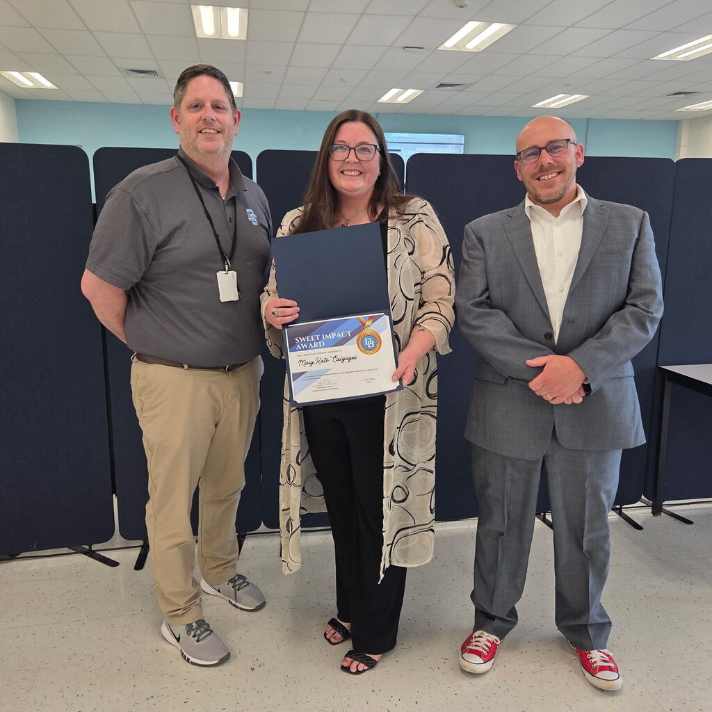 Principal Jim Towers standing with English Teacher Mary Kate Calcagni and Superintendent Tom Voelker. Mary Kate is holding a Sweet Impact Award Certificate. The trio are standing in the Daniel Boone Board Room.