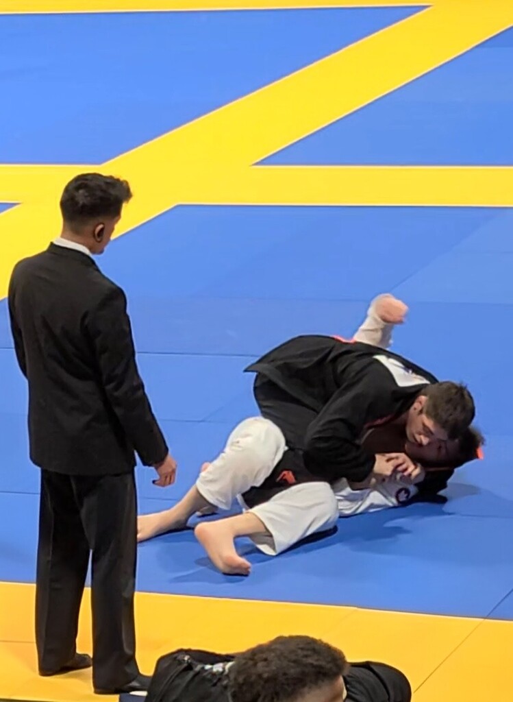 Two jiu-jitsu competitors competing on the mat while a man in a black suit watches.