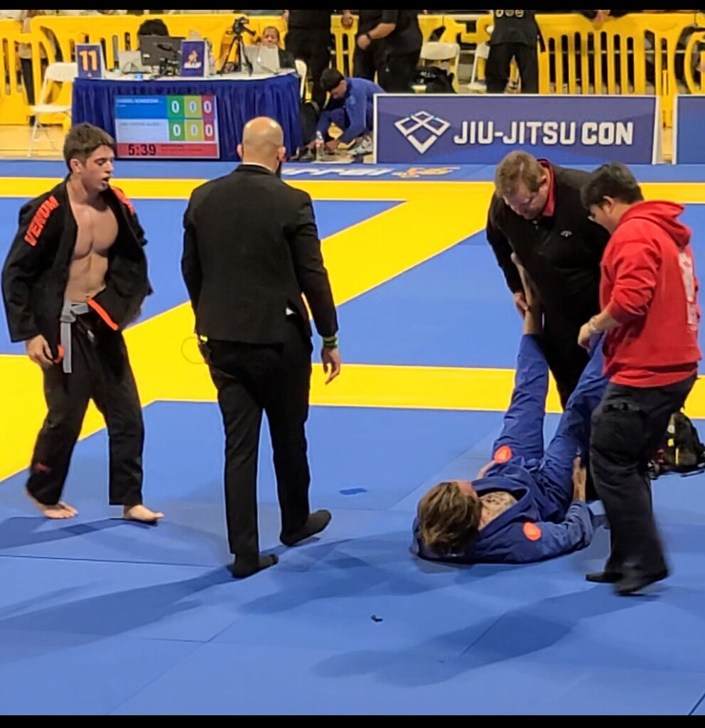 One jiu-jitsu competitor standing watching two helpers attending to an opponent laying on the mat. An official in a black suit with his back to the camera walks between them.