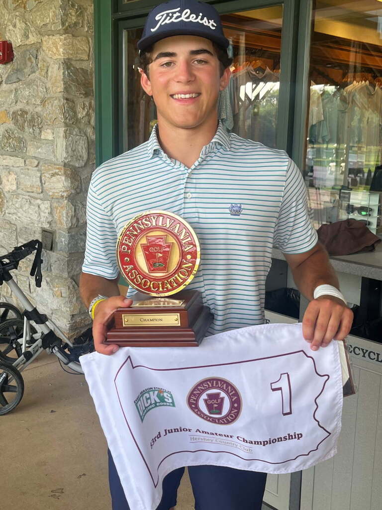 A smiling young golfer stands outside the Hershey Country Club clubhouse holding the Pennsylvania Golf Association Junior Amateur Champion trophy and the championship flag. He is wearing a navy Titleist hat, a light blue and white striped polo shirt, and navy pants. The white championship flag features the Pennsylvania outline, Pennsylvania Golf Association logo, and a large number 1, recognizing his first-place finish at the 53rd Pennsylvania Junior Amateur Championship.
