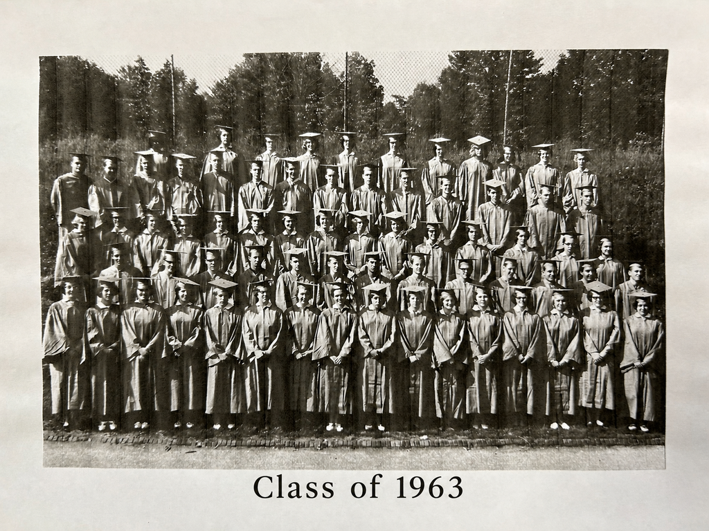 Black-and-white archival class photo labeled "Class of 1963" at the bottom. Dozens of graduating seniors wearing caps and gowns are arranged in several rows outdoors, standing in front of a grassy area with tall trees and a chain-link fence in the background. The vintage photograph has the look of a yearbook or graduation keepsake and is framed by a white border.
