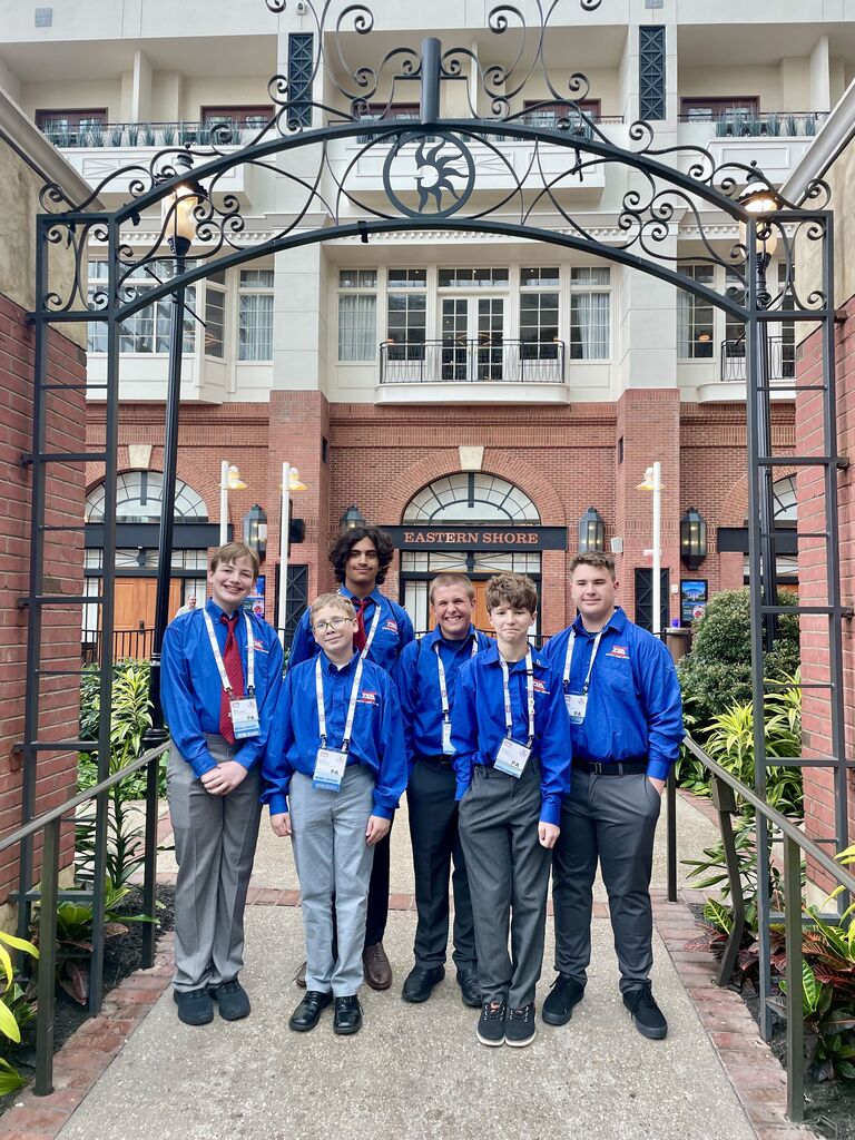 Six Daniel Boone TSA students pose beneath a decorative wrought-iron archway outside a hotel in Washington, D.C. The students wear matching blue TSA shirts and conference badges as they smile together in a landscaped courtyard.