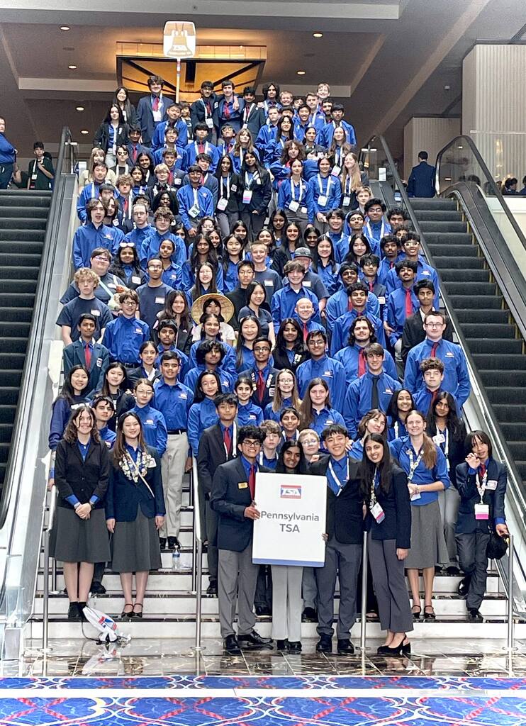 A large group of Pennsylvania Technology Student Association (TSA) students and advisors gather on a grand staircase inside the National TSA Conference venue. Participants wear blue TSA shirts, conference attire, and name badges, with students in the front holding a sign reading "Pennsylvania TSA."