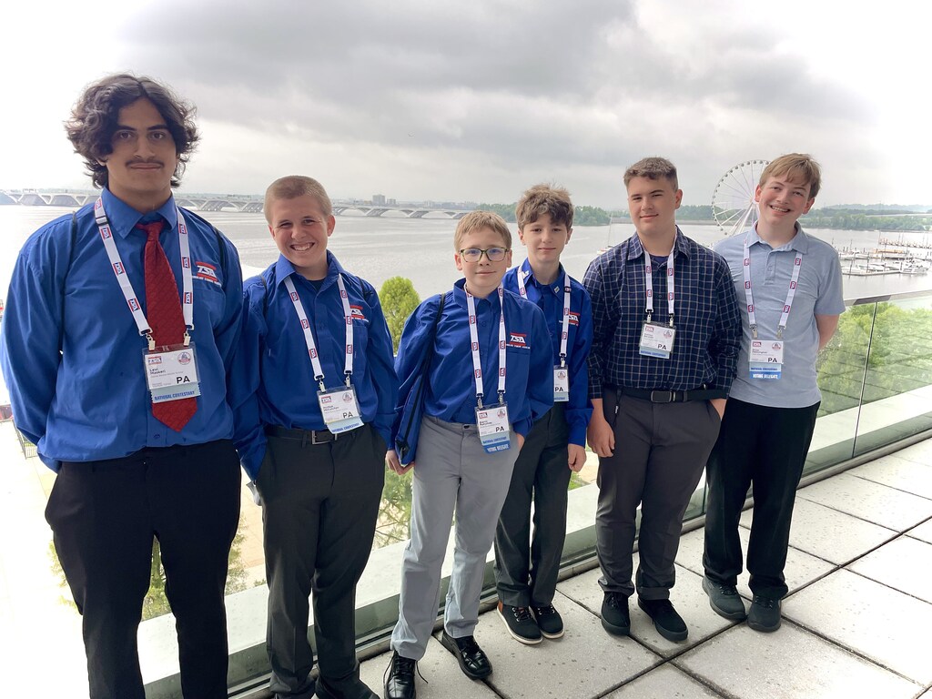 Six Daniel Boone Technology Student Association (TSA) students stand together on an outdoor balcony overlooking the Washington, D.C., waterfront. Wearing TSA conference badges and blue TSA shirts, they smile for the camera with a river, bridge, Ferris wheel, and cloudy sky in the background.