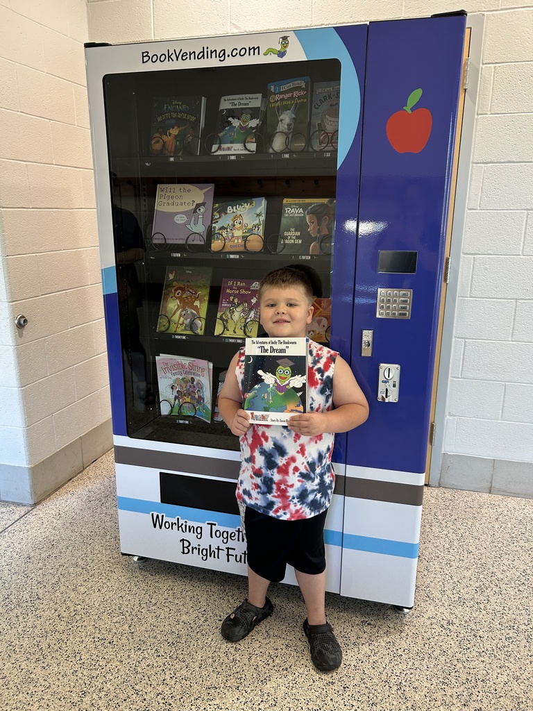 One student standing in front of a book vending machine. Student his holding a Inchy the Bookworm book titled "The Dream".